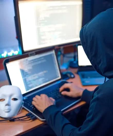 Vista de costas de uma pessoa com capuz escuro digitando em um notebook com códigos na tela, em um ambiente de hacker com múltiplos monitores e uma máscara branca sobre a mesa. Dicas de segurança para se proteger de ataques hacker