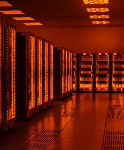 Fotografia em perspectiva de um corredor de data center moderno, com várias fileiras de racks de servidores pretos iluminados por uma intensa e brilhante luz laranja e âmbar, refletindo no chão e no teto.