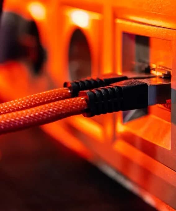 Dois cabos de rede trançados de cor vermelha com conectores pretos, conectados lado a lado em tomadas de parede RJ45. A cena é iluminada por uma luz laranja vibrante que destaca os cabos em primeiro plano, mantendo o fundo levemente desfocado. Internet 100% Fibra em Londrina