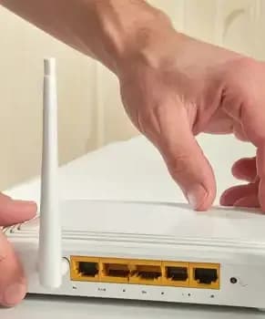 Mãos de um homem ajustando ou configurando um roteador Wi-Fi branco com duas antenas sobre uma mesa clara, mostrando o painel traseiro com portas de rede LAN amarelas. Como testar a velocidade da internet e entender o resultado?