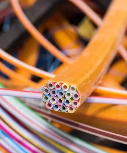 Vista em primeiro plano do corte transversal de um cabo de fibra óptica laranja, revelando vários microdutos coloridos em seu interior, com outros cabos de rede desfocados ao fundo. 4 cuidados com a fibra óptica