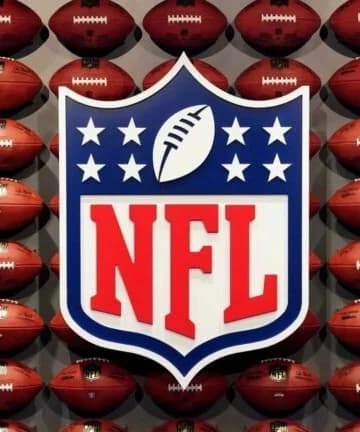 O logotipo oficial da NFL em formato de escudo destaca-se no centro da imagem com letras vermelhas em negrito, posicionado à frente de uma parede inteiramente coberta por bolas de futebol americano de couro marrom organizadas em fileiras simétricas.